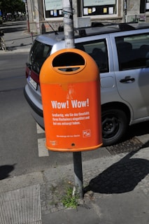 A bright orange public trash bin is mounted on a pole next to a parked car on a city street. The bin has text printed on it, promoting responsible waste disposal, specifically about wrapping and disposing of pet waste. The background shows part of a building with windows and a cyclist in the distance.