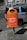 A bright orange public trash bin is mounted on a pole next to a parked car on a city street. The bin has text printed on it, promoting responsible waste disposal, specifically about wrapping and disposing of pet waste. The background shows part of a building with windows and a cyclist in the distance.