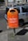 A bright orange public trash bin is mounted on a pole next to a parked car on a city street. The bin has text printed on it, promoting responsible waste disposal, specifically about wrapping and disposing of pet waste. The background shows part of a building with windows and a cyclist in the distance.