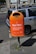 A bright orange public trash bin is mounted on a pole next to a parked car on a city street. The bin has text printed on it, promoting responsible waste disposal, specifically about wrapping and disposing of pet waste. The background shows part of a building with windows and a cyclist in the distance.