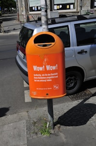 A bright orange public trash bin is mounted on a pole next to a parked car on a city street. The bin has text printed on it, promoting responsible waste disposal, specifically about wrapping and disposing of pet waste. The background shows part of a building with windows and a cyclist in the distance.