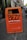 An orange public trash bin is mounted on a pole along a city sidewalk. The bin has graffiti spray-painted across it, with text promoting proper disposal of dog waste below. The surrounding area includes a cobblestone path and parked cars, with buildings lining the street.