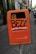 An orange public trash bin is mounted on a pole along a city sidewalk. The bin has graffiti spray-painted across it, with text promoting proper disposal of dog waste below. The surrounding area includes a cobblestone path and parked cars, with buildings lining the street.