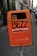 An orange public trash bin is mounted on a pole along a city sidewalk. The bin has graffiti spray-painted across it, with text promoting proper disposal of dog waste below. The surrounding area includes a cobblestone path and parked cars, with buildings lining the street.