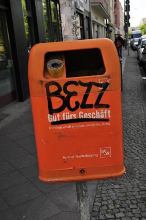 An orange public trash bin is mounted on a pole along a city sidewalk. The bin has graffiti spray-painted across it, with text promoting proper disposal of dog waste below. The surrounding area includes a cobblestone path and parked cars, with buildings lining the street.