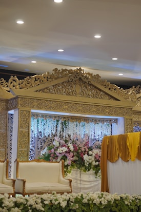 A richly decorated stage setup featuring ornate gold carvings and detailed floral arrangements. The backdrop showcases a combination of white and pastel pink flowers, adding an elegant touch. Draped fabric in a mustard hue hangs to the right, complementing the luxurious feel. White upholstered chairs with wooden frames are placed in front, surrounded by a border of white flowers.