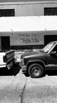 A black and white photo capturing two parked trucks close to a concrete building. The building has graffiti text written in Spanish on the wall, stating, 'Dices ser libre... tengo mis dudas...'. The overall scene is urban and appears to be taken on a quiet street.