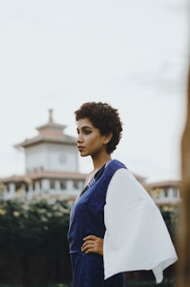 A person stands in profile against a blurred background of a building with traditional architecture. The person has short curly hair and is wearing a blue outfit with a white sleeve. Their posture is confident as they look into the distance.