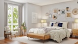 A beautifully decorated bedroom features a large bed with white and beige linens, accented by dark blue and patterned pillows. Above the bed is an arrangement of various framed artworks and decorative plates on the wall. A wooden bench sits at the foot of the bed. To the left, a cozy armchair with a pillow is placed beside a window with long, light-colored curtains allowing natural light to brighten the room. Beside the chair, a small round side table holds a couple of books and a plant. Another plant in a decorative pot is positioned next to a wooden bedside table, complemented by a modern lamp.