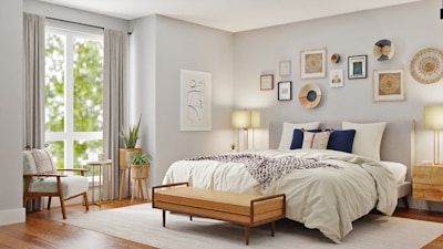 A beautifully decorated bedroom features a large bed with white and beige linens, accented by dark blue and patterned pillows. Above the bed is an arrangement of various framed artworks and decorative plates on the wall. A wooden bench sits at the foot of the bed. To the left, a cozy armchair with a pillow is placed beside a window with long, light-colored curtains allowing natural light to brighten the room. Beside the chair, a small round side table holds a couple of books and a plant. Another plant in a decorative pot is positioned next to a wooden bedside table, complemented by a modern lamp.