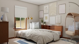 A cozy bedroom features a neatly made bed with light gray bedding, adjacent to a wooden nightstand with a gold lamp. Above the bed, an arrangement of abstract and minimalist art decorates the wall. A window with white blinds allows natural light into the room. In the corner, a wicker hanging chair equipped with cushions adds a stylish touch. Neutral tones dominate the space, with hints of blue in the rug and decor items.