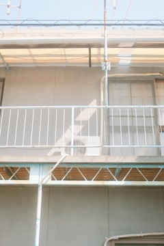 A section of a building facade with a small balcony featuring a white metal railing. The balcony has an air conditioning unit partially visible. Overhead, there is a translucent awning supported by metal frames. The structure appears to be undergoing some form of maintenance or renovation, indicated by scaffolding present around the balcony.