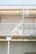 A section of a building facade with a small balcony featuring a white metal railing. The balcony has an air conditioning unit partially visible. Overhead, there is a translucent awning supported by metal frames. The structure appears to be undergoing some form of maintenance or renovation, indicated by scaffolding present around the balcony.
