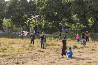 A group of people is gathered in an open field with trees in the background, engaging in kite flying activities. Adults and children are present, with several colorful kites in the sky. The scene is lively with people interacting and enjoying the outdoor activity.
