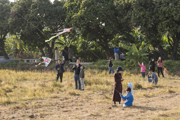 A group of people is gathered in an open field with trees in the background, engaging in kite flying activities. Adults and children are present, with several colorful kites in the sky. The scene is lively with people interacting and enjoying the outdoor activity.