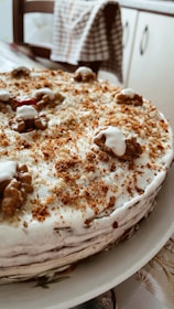 A homemade cake topped with a layer of white frosting and sprinkled with brown crumbs. Walnuts are placed decoratively on top of the cake. The cake is positioned on a white plate in a cozy kitchen setting.