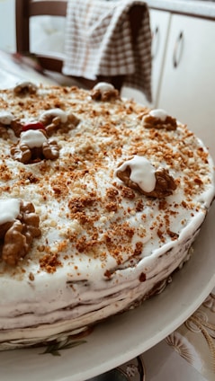 A homemade cake topped with a layer of white frosting and sprinkled with brown crumbs. Walnuts are placed decoratively on top of the cake. The cake is positioned on a white plate in a cozy kitchen setting.