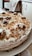 A homemade cake topped with a layer of white frosting and sprinkled with brown crumbs. Walnuts are placed decoratively on top of the cake. The cake is positioned on a white plate in a cozy kitchen setting.