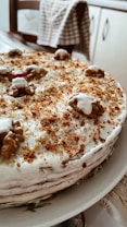A homemade cake topped with a layer of white frosting and sprinkled with brown crumbs. Walnuts are placed decoratively on top of the cake. The cake is positioned on a white plate in a cozy kitchen setting.