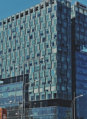 A modern glass office building with a geometric pattern of windows, reflecting the blue sky. Streetlights line the sidewalk below, and part of a billboard is visible in the foreground.