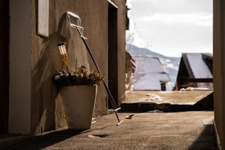 A narrow alleyway with a snow-covered landscape in the background. A white planter with plants and a solar torch is leaning against a wall, alongside a purple snow shovel. The ground is partially covered with melting snow, and the scene is illuminated by soft sunlight casting shadows.