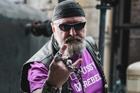 A man with a beard is wearing a black bandana, sunglasses, and a leather vest over a purple shirt that features white text. He has a defiant expression and holds up his hand with his fingers making a rock-and-roll symbol while wearing multiple rings and bracelets. The background is industrial, with blurred machinery and structures.