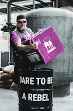 A person with a rugged appearance, wearing sunglasses and a headband, stands holding a purple box labeled 'PETREBELS' over a black barrel. The barrel has the text 'DARE TO BE A REBEL' written on it. The scene is set in an industrial environment with metal pipes and a large tank in the background.