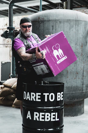 A person with a rugged appearance, wearing sunglasses and a headband, stands holding a purple box labeled 'PETREBELS' over a black barrel. The barrel has the text 'DARE TO BE A REBEL' written on it. The scene is set in an industrial environment with metal pipes and a large tank in the background.