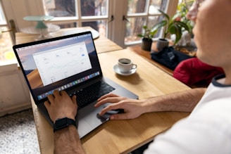 A person is working on a laptop at a wooden table, with charts and graphs displayed on the screen. Nearby, there is a cup of coffee, a potted plant, and some folders or bags on the table. The scene is well-lit by natural light coming through large windows in the background.
