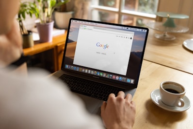A person is using a laptop with the Google homepage displayed on the screen. The setting is a cozy room with natural light coming in through a window. There is a warm cup of coffee on the table next to the laptop, and potted plants in the background.