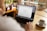 A person is using a laptop with the Google homepage displayed on the screen. The setting is a cozy room with natural light coming in through a window. There is a warm cup of coffee on the table next to the laptop, and potted plants in the background.