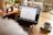 A person is using a laptop with the Google homepage displayed on the screen. The setting is a cozy room with natural light coming in through a window. There is a warm cup of coffee on the table next to the laptop, and potted plants in the background.