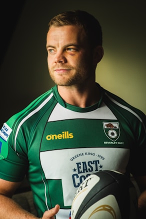 A man, wearing a green and white rugby jersey with the text 'Beverley RUFC' and 'oneills' in yellow on it, holds a rugby ball. He appears to have a focused or determined expression. The background is dark with a light source creating a soft shadow on his face.