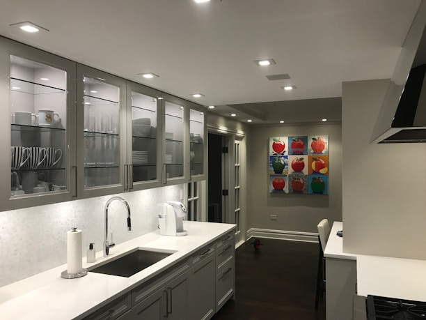 A modern kitchen with a sleek design featuring white and grey tones. Cabinets with glass doors display neatly arranged dishes and cups. The countertop holds a paper towel roll, a coffee maker, and a sink with a shiny faucet. The walls are decorated with colorful pop art paintings depicting fruits. Recessed ceiling lights illuminate the space.