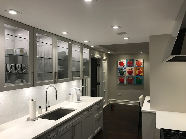 A modern kitchen with a sleek design featuring white and grey tones. Cabinets with glass doors display neatly arranged dishes and cups. The countertop holds a paper towel roll, a coffee maker, and a sink with a shiny faucet. The walls are decorated with colorful pop art paintings depicting fruits. Recessed ceiling lights illuminate the space.