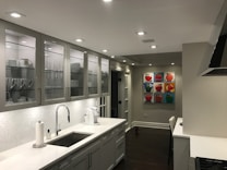 A modern kitchen with a sleek design featuring white and grey tones. Cabinets with glass doors display neatly arranged dishes and cups. The countertop holds a paper towel roll, a coffee maker, and a sink with a shiny faucet. The walls are decorated with colorful pop art paintings depicting fruits. Recessed ceiling lights illuminate the space.