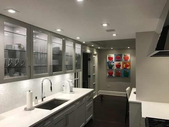 A modern kitchen with a sleek design featuring white and grey tones. Cabinets with glass doors display neatly arranged dishes and cups. The countertop holds a paper towel roll, a coffee maker, and a sink with a shiny faucet. The walls are decorated with colorful pop art paintings depicting fruits. Recessed ceiling lights illuminate the space.