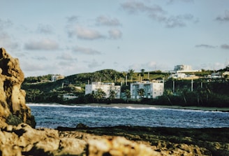 A coastal landscape features rocky foreground with waves crashing against it. In the middle ground, there is a sandy shoreline and an array of buildings surrounded by lush greenery. The background shows a hill with scattered buildings under a partly cloudy sky.