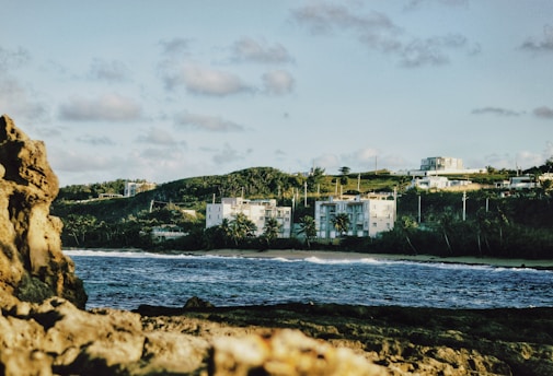 A coastal landscape features rocky foreground with waves crashing against it. In the middle ground, there is a sandy shoreline and an array of buildings surrounded by lush greenery. The background shows a hill with scattered buildings under a partly cloudy sky.