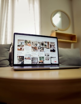 A laptop displaying a webpage with several interior design images is placed on a wooden table. Behind it, a gray couch and a round mirror on the wall can be seen, suggesting a cozy room setup. Soft lighting filters through sheer curtains in the background.