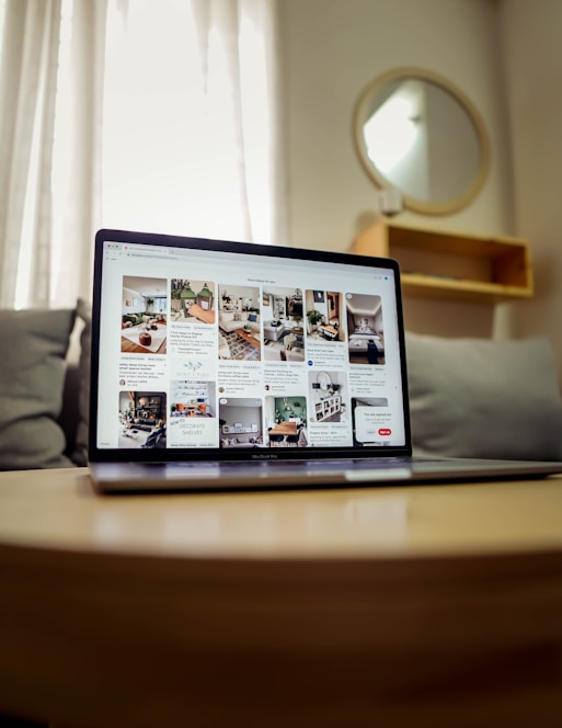 A laptop displaying a webpage with several interior design images is placed on a wooden table. Behind it, a gray couch and a round mirror on the wall can be seen, suggesting a cozy room setup. Soft lighting filters through sheer curtains in the background.