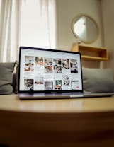 A laptop displaying a webpage with several interior design images is placed on a wooden table. Behind it, a gray couch and a round mirror on the wall can be seen, suggesting a cozy room setup. Soft lighting filters through sheer curtains in the background.