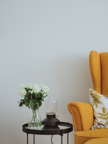 A modern and minimalist interior setup featuring a mustard-yellow armchair with a decorative pillow and a side table. The table holds a vase filled with white roses and a small decorative lamp with a glass cover. The background is a plain light-colored wall, contributing to a serene and elegant ambiance.