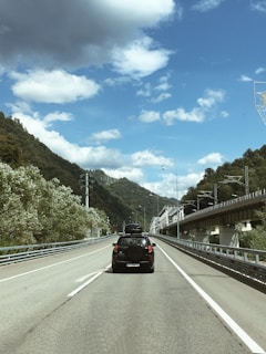 A mountain road with a black SUV driving ahead. The vehicle has a roof carrier and spare tire at the back. On either side, lush green hills rise up, and a railway bridge runs parallel to the road on the right. The sky is partly cloudy, featuring a mix of white fluffy clouds and some dark grey clouds.