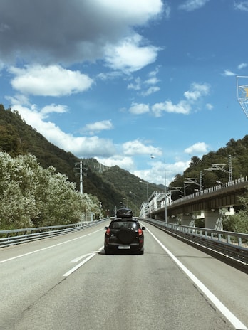 A mountain road with a black SUV driving ahead. The vehicle has a roof carrier and spare tire at the back. On either side, lush green hills rise up, and a railway bridge runs parallel to the road on the right. The sky is partly cloudy, featuring a mix of white fluffy clouds and some dark grey clouds.
