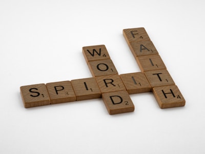 Scrabble tiles arranged in a crossword puzzle layout, featuring intersecting words like 'WORD,' 'SPIRIT,' and 'FAITH.' The tiles are light brown with black letters and numbers indicating the point value for each letter.
