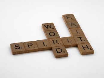 Scrabble tiles arranged in a crossword puzzle layout, featuring intersecting words like 'WORD,' 'SPIRIT,' and 'FAITH.' The tiles are light brown with black letters and numbers indicating the point value for each letter.