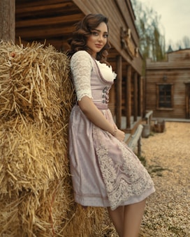 A woman in a traditional dress with intricate lace patterns stands leaning against a stack of hay. Her curly hair is styled elegantly, and she's positioned near a rustic wooden building on a gravel path. The background suggests a rural, possibly historical setting with overcast skies.