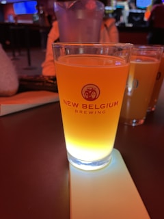 A glass filled with an amber-colored beverage from New Belgium Brewing, sitting on a coaster. The scene takes place in a dimly lit bar, with additional glasses visible in the background.