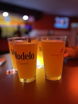 Two glasses of beer are placed on a dimly lit tabletop, with one glass labeled 'Modelo Especial' and the other 'New Belgium Brewing'. The background is blurred, showing warm, ambient lighting and hints of a bar or social setting.